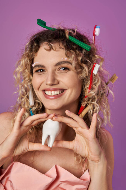 sorridente mulher loira com escovas de dentes em seu cabelo encaracolado segurando dente, conceito de fada do dente - Foto, Imagem