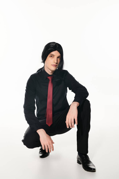 homme à la mode en vêtements noirs et perruque avec cravate rouge assis sur des hanches sur blanc, tendance cosplay - Photo, image