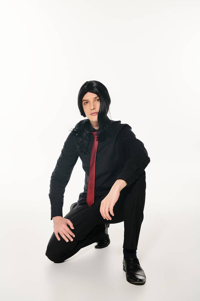 cosplay man in black clothes and wig with red tie posing on haunches looking at camera on white - Photo, Image