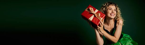 Yeşil elbiseli mutlu yeni yıl elfi elinde hediyeyle poz veriyor. Koyu yeşil arka plan, pankart. - Fotoğraf, Görsel