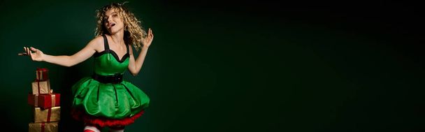 χαρούμενη γυναίκα με κοστούμι ξωτικό gesturing με τα χέρια που θέτουν κοντά σωρός από δώρα σε πράσινο φόντο, πανό - Φωτογραφία, εικόνα