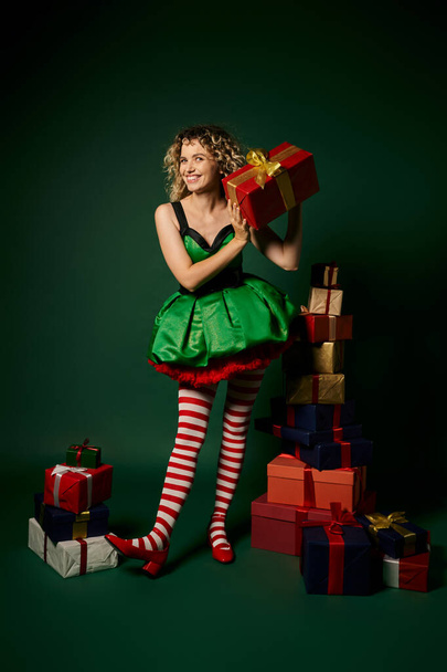 curly new year elf standing near pile of gifts and mysteriously showing one of them at camera - Photo, Image