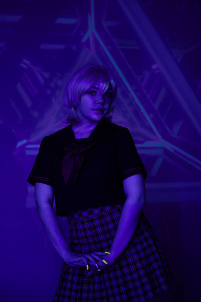 young blonde woman in school uniform in neon light on blue abstract backdrop, cosplay character - Photo, Image