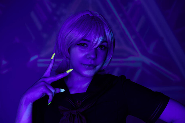 mujer de estilo anime con peluca rubia y manicura fluorescente mostrando signo de victoria en luz de neón azul - Foto, imagen