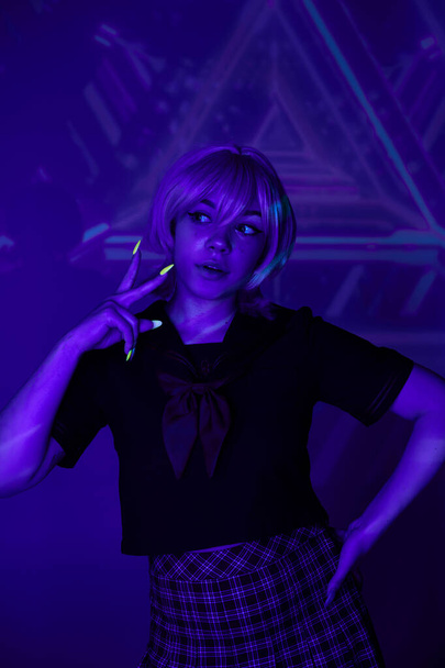 femme blonde en uniforme scolaire avec la main sur la hanche montrant signe de victoire dans la lumière néon bleu, style anime - Photo, image