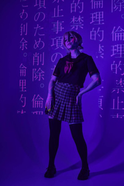 mulher estilo anime em uniforme escolar com a mão no quadril em luz de néon azul com projeção hieróglifos - Foto, Imagem