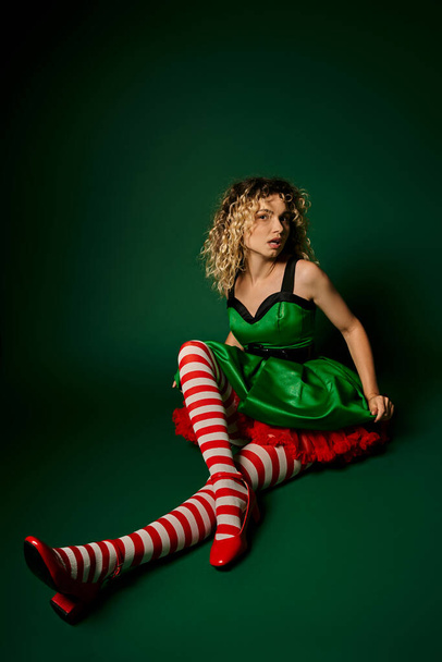 dreamy curly woman dressed as new year elf sitting on floor posing on dark green background - Photo, Image