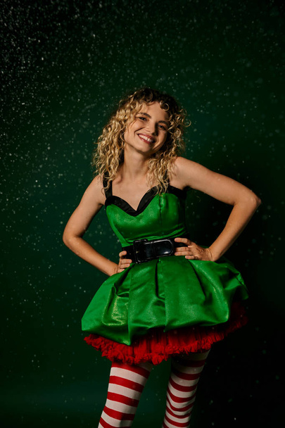 cheerful woman in green festive dress smiling and holding hands akimbo, new year elf concept - Photo, Image