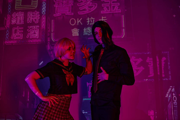 young woman in school uniform scaring anime style boyfriend on purple neon backdrop with hieroglyphs - Photo, Image
