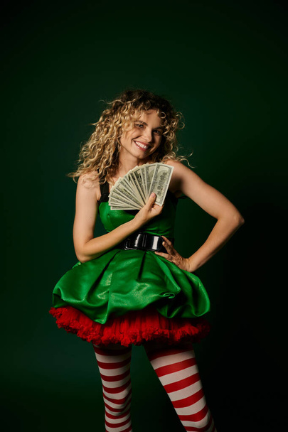 smiley krullende elf in groen nieuwjaar jurk en gestreepte kousen met contant geld met een arm akimbo - Foto, afbeelding