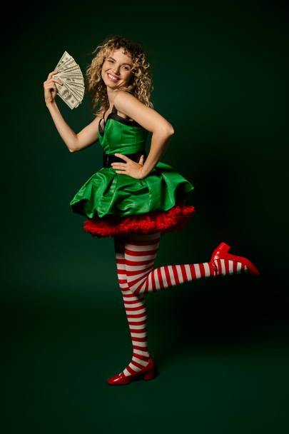docela nový rok elf stojí na jedné noze drží peníze a usmívá se na kameru s jednou rukou akimbo - Fotografie, Obrázek