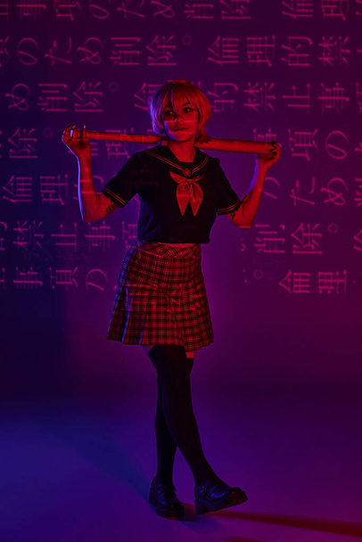 trendy cosplay woman in school uniform with baseball bat on neon purple backdrop with hieroglyphs - Photo, Image