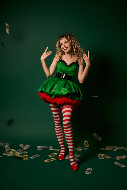 smiley new year elf in green dress and stockings gesturing and smiling with cash on green floor - Photo, Image