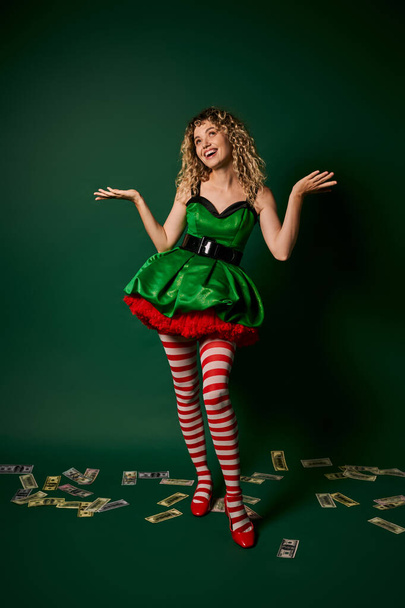 cheerful woman in green dress looking up and gesturing with money on floor, new year elf concept - Photo, Image