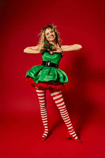 atractiva mujer vestida como elfo año nuevo sonriendo y sosteniendo presente en la cámara en el telón de fondo rojo - Foto, imagen