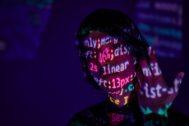 Studentin im Anime-Stil mit schwarzer Perücke in Neonlicht mit Programmiersymbolen auf dunkelblauem Hintergrund - Foto, Bild