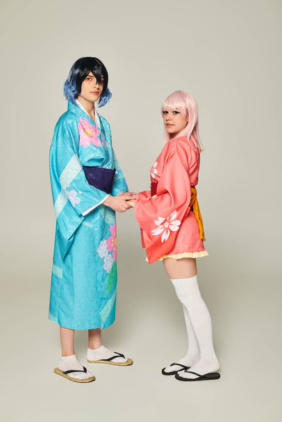 pareja joven en coloridos kimonos tomados de la mano y mirando a la cámara en gris, concepto de anime - Foto, Imagen