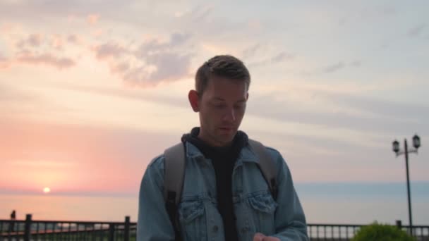 Jonge knappe man reiziger met behulp van smartphone voor boek accommodatie buiten in de buurt van de zeekust bij zonsondergang. Blanke man browsen toepassingen op zijn mobiele telefoon op zoek naar overnachting. Zomer - Video