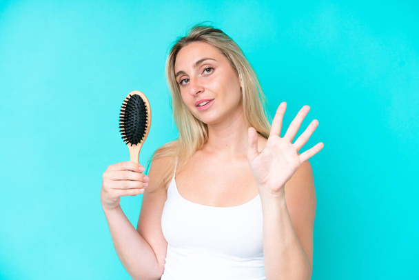 Young caucasian woman with hair comb isolated on blue background saluting with hand with happy expression - Foto, Imagen