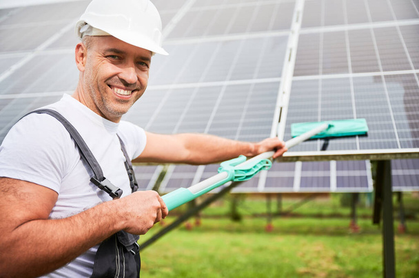 Usmívající se muž čistí fotovoltaickou baterii mop. Profesionální pracovník odvádí dobrou práci při čištění solárního panelu od prachu. Muž držící mop a usmívající se přímo do kamery. - Fotografie, Obrázek