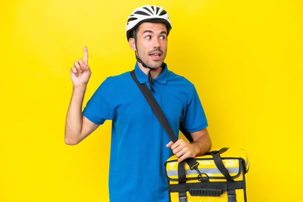 Young handsome man with thermal backpack over isolated yellow background intending to realizes the solution while lifting a finger up - Photo, Image