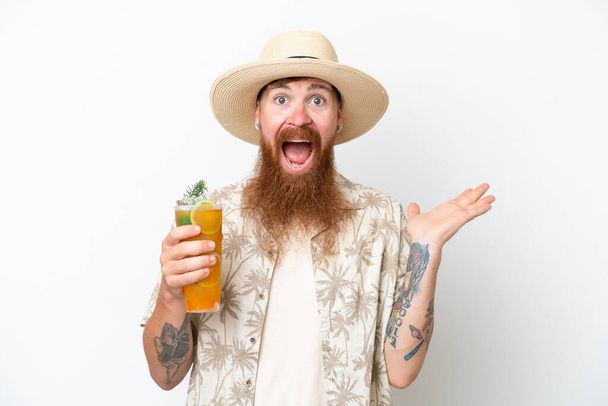 Uomo dai capelli rossi con lunga barba che beve un cocktail su una spiaggia isolata su sfondo bianco con espressione facciale scioccata - Foto, immagini