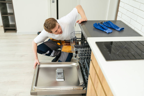 Technician or worker in uniform installs dishwasher into the kitchen furniture. Repairman wear worker suit repairing maintenance of dishwasher. Master in protective gloves fix dishwasher. - Photo, Image