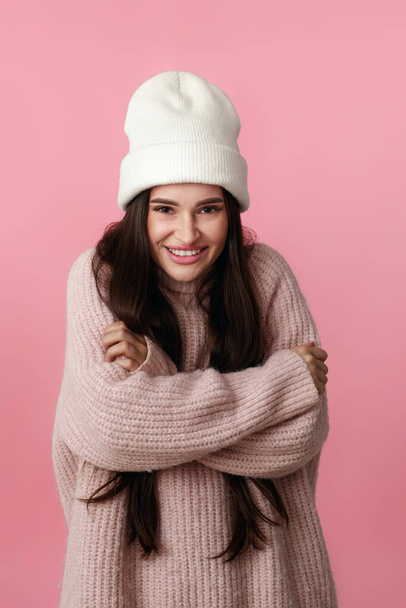 pretty woman in white hat, sweater hugging herself and smiling feeling happy on pastel pink background. - Foto, Bild