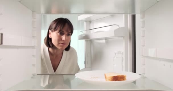 Tyytymätön nainen tarkistaa voileivän tuoreuden jääkaapissa ja valitsee sitten vesipullon. Tyytymätön ilme viittaa siihen, ettei hän ehkä pidä voileivästä.. - Materiaali, video