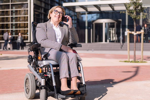 smiling businesswoman using wheelchair talking by mobile phone in front of an office building in the financial district, concept of diversity and technology of communication - Фото, изображение