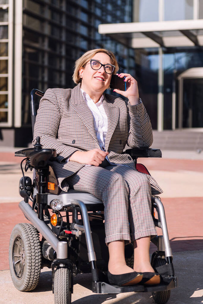 smiling businesswoman using wheelchair talking by mobile phone in front of an office building in the financial district, concept of diversity and technology of communication - Foto, afbeelding