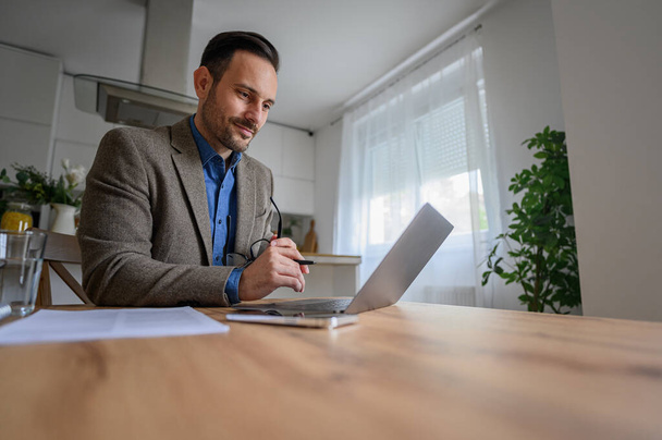 Professionista uomo sicuro in giacca e cravatta guardando il computer wireless sulla scrivania mentre si lavora a casa - Foto, immagini