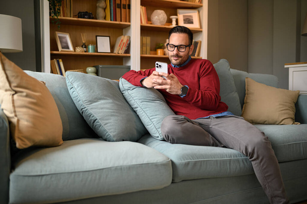 Fešák mladý muž procházení sociálních médií na chytrém telefonu při relaxaci na pohovce v obývacím pokoji - Fotografie, Obrázek