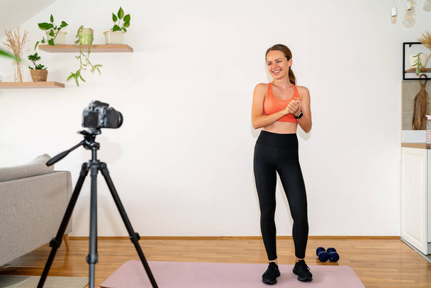 Νεαρή αθλήτρια γυμναστικής φορώντας αθλητικά να στέκεται μπροστά στην κάμερα στο τρίποδο και να καταγράφει το βίντεο μπλογκ της για τον υγιεινό τρόπο ζωής. - Φωτογραφία, εικόνα