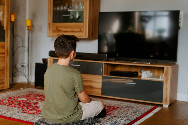 Zadní pohled na nerozeznatelné dítě v neformálním oblečení sedí na podlaze se zkříženýma nohama a dívá se na televizi - Fotografie, Obrázek