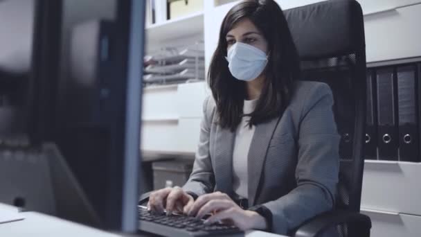 Jeune spécialiste confiante en costume formel et masque de protection assis au bureau et utilisant un ordinateur tout en travaillant dans un espace de travail moderne - Séquence, vidéo