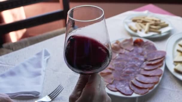 Zbliżenie upraw nierozpoznawalny starszy człowiek z pomarszczonymi rękami mieszając czerwone wino w szklance w zwolnionym tempie siedząc przy stole z przekąskami w restauracji - Materiał filmowy, wideo