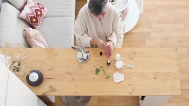 Desde arriba de la mujer de mediana edad en bata de baño goteando suero en la mano mientras está sentado en la mesa durante la rutina de cuidado de la piel en casa - Metraje, vídeo