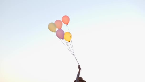 W czasie rzeczywistym optymistyczna kobieta w kapeluszu tańczy z kolorowymi balonami stojąc na brzegu w pobliżu falującego jeziora podczas zachodu słońca - Materiał filmowy, wideo