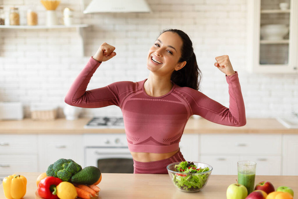 El poder de la comida sana. Happy Young Fitness Lady mostrando músculos bíceps mientras cocina ensalada, perdiendo peso final disfrutando de la cena, de pie cerca de la mesa de la cocina con verduras en casa, sonriendo a la cámara - Foto, Imagen