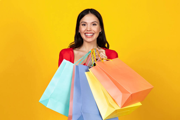 Porträt einer glücklichen schönen Frau, die helle Einkaufstaschen in den Händen hält und in die Kamera blickt, lächelnde Shopaholic Lady, die saisonale Verkäufe genießt, vor gelbem Studiohintergrund posiert, Kopierraum - Foto, Bild