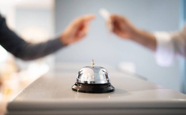 Call Bell Standing On Reception Counter At Hotel Lobby While Receptionist Giving Key To Tourist Man, Selective Focus, Closeup. Guest Checks In At Accommodation, Takes Room Keys From Hostel Staff - Photo, Image