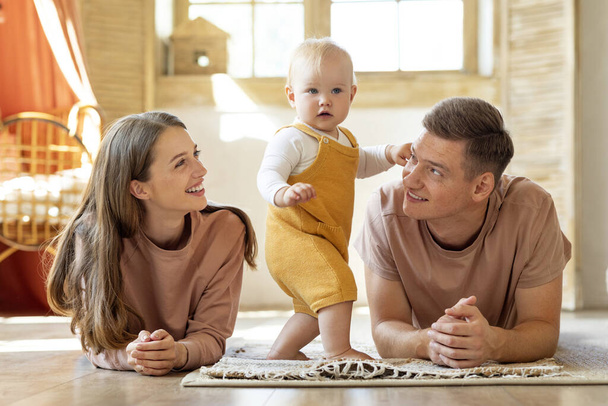 Счастливые молодые родители, глядя на своего прелестного малыша, пока они отдыхают вместе дома, улыбаются маме и папе, сближаясь с младенцем во время отдыха на полу в гостиной, бесплатное пространство - Фото, изображение