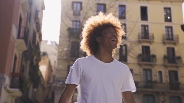 Alhaalta käsin ammuttu iloinen nuori afroamerikkalainen mies valkoisessa t-paidassa, jossa afro-kampaus hymyilee ja katsoo pois kävellessään kaupungin kadulla rakennusten lähellä. - Materiaali, video