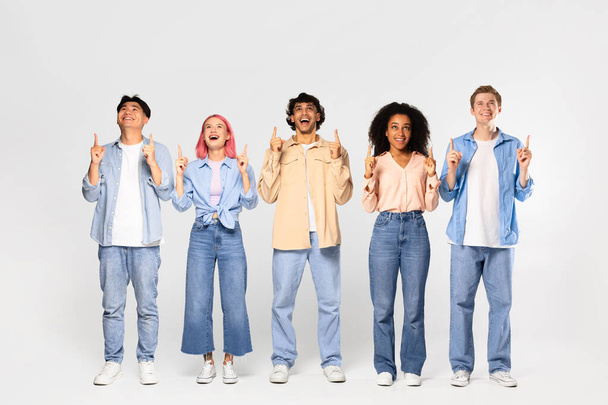 Group of young excited diverse friends looking up with excitement and pointing fingers up at copy space standing together over white background, full length shot - Photo, Image