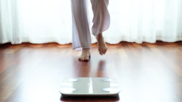 Top view crop faceless barefoot female in cozy pajama standing on digital weight and body fat scales with display showing healthy weight of 60 kg on bathroom floor in morning - Footage, Video