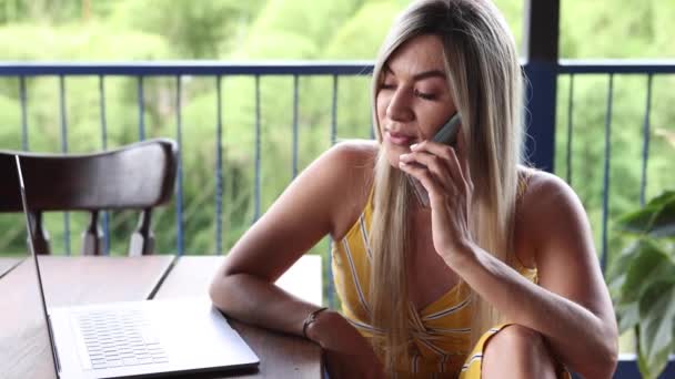Mujer joven en vestido de verano casual netbook de navegación mientras habla por teléfono en la terraza del restaurante al aire libre - Metraje, vídeo