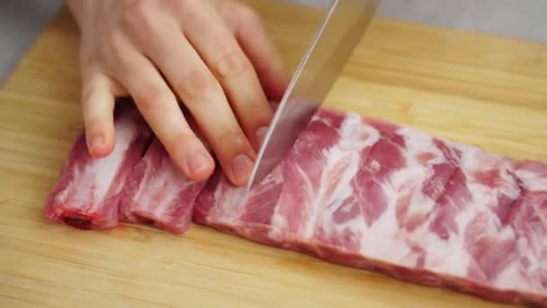 Tracking shot van anonieme vrouw snijden van verse varkensribben met scherp mes op houten snijplank tijdens de bereiding van voedsel in de keuken - Video