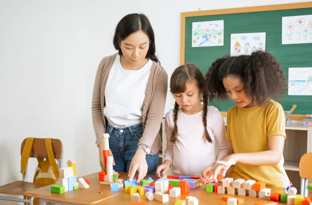 Многонациональная группа детей, стоящих и играющих с красочными блоками на столе с учительницей-азиаткой в классе. Развитие мозговой подготовки для развития навыков детей. - Фото, изображение