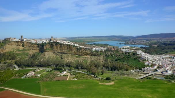 Links Drohnenblick auf grüne Farmfelder in der Nähe der Klippe mit der Stadt Arcos de la Frontera vor wolkenlosem blauem Himmel in Andalusien, Spanien - Filmmaterial, Video